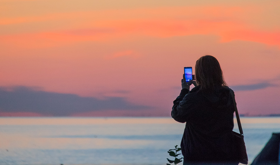 A woman holds up a phone to take a photo of a sunset. 