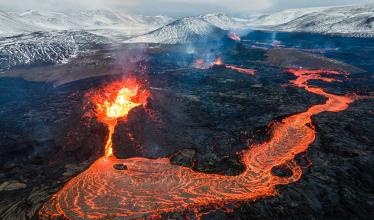 Glowing lava flows in the foreground with snow capped volcanic mountains in the background.