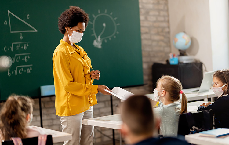 A teacher, wearing a mask, stands near a student, also wearing a mask, in a classroom 