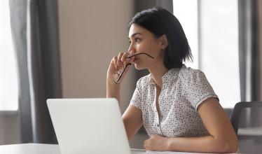 Woman holding glasses to mouth, gazing over laptop. 