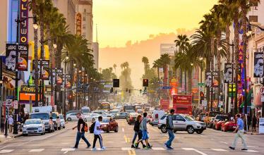 Golden sunlight bathes Los Angeles street, with cars, pedestrians, marquees, and some trees.