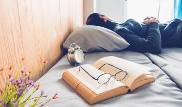 Man sleeps on a bed, with a book in the foreground, glasses on top of it, with an alarm clock nearby.